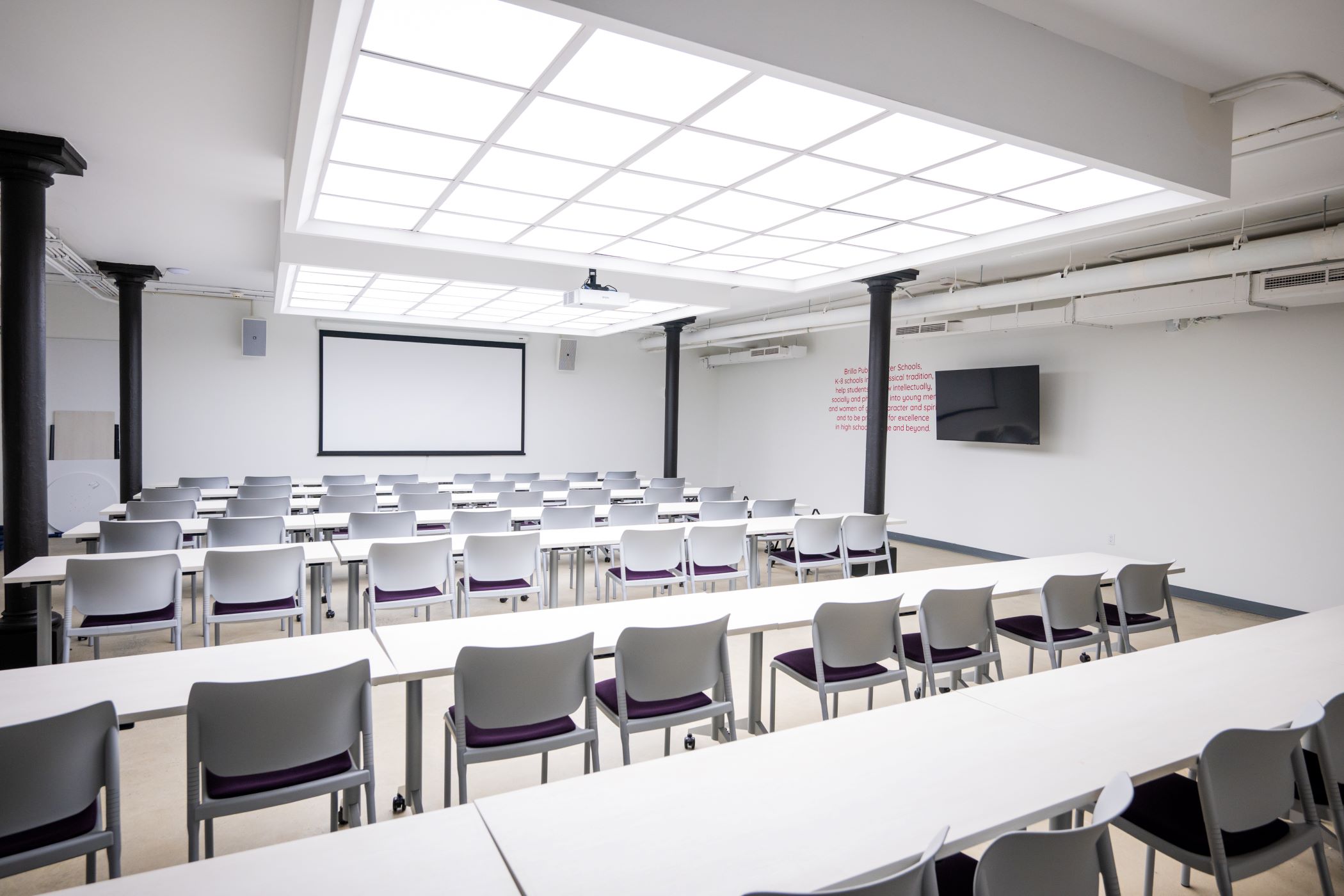 artificially lit white room with rows of tables and chairs facing a projector screen