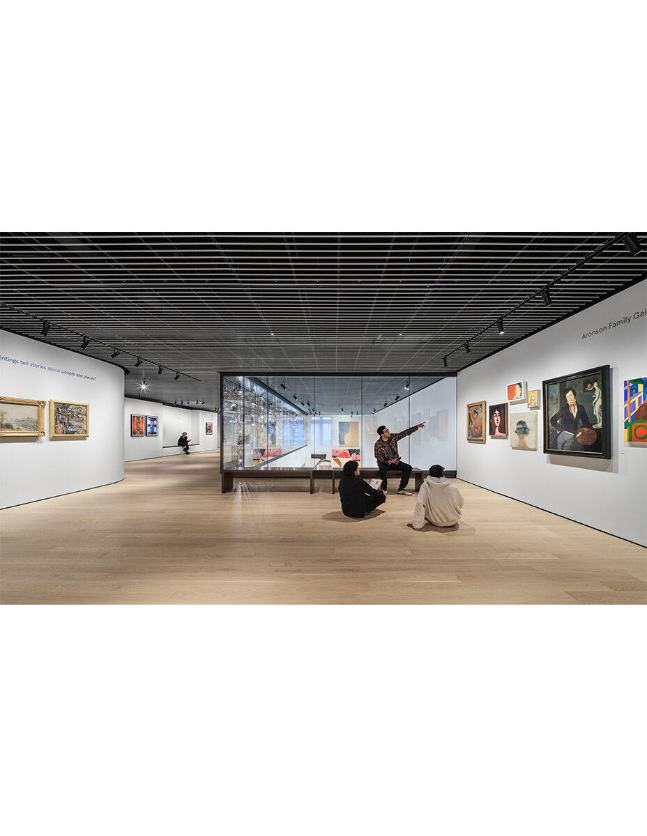 Museum gallery with clean white walls, light wood floors, dark striped ceiling and glass opening to space below