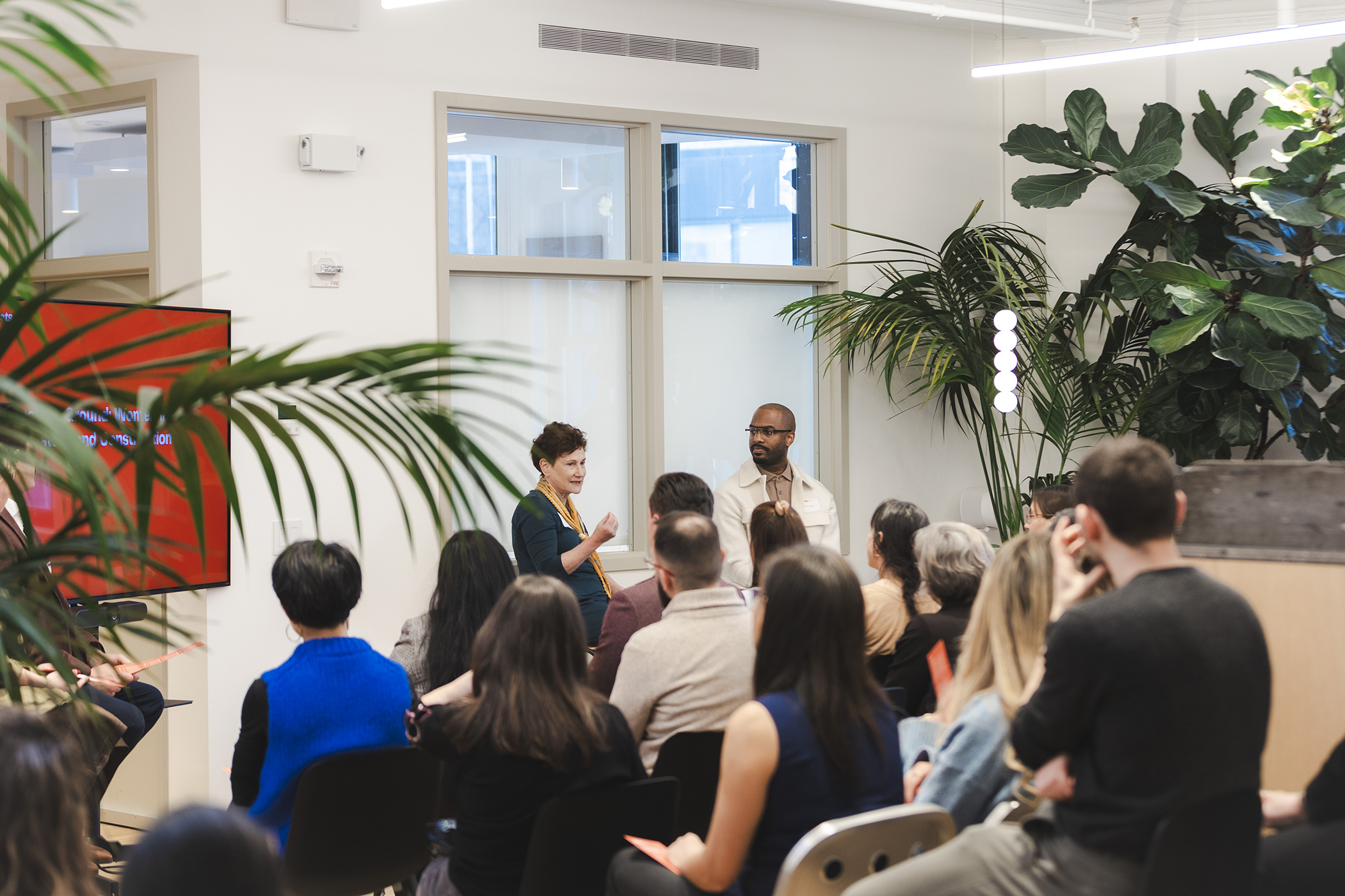A woman and a man speaking in front of a small seated crowd gathered in a plant-filled office
